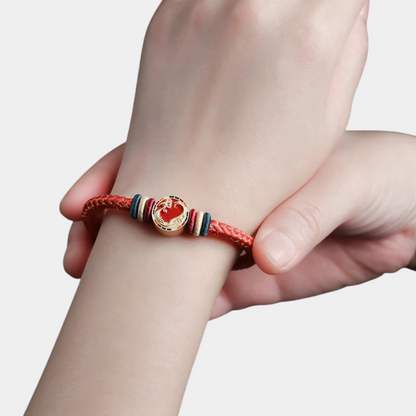 Hand wearing a red braided bracelet with a heart charm on a light gray background